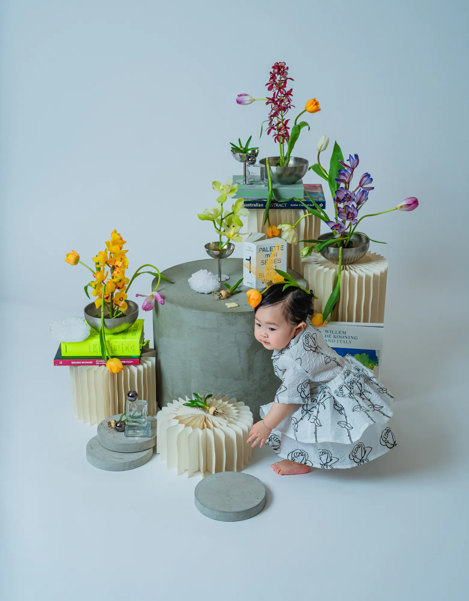 花と本のアート什器に手を伸ばす白いワンピースの赤ちゃん