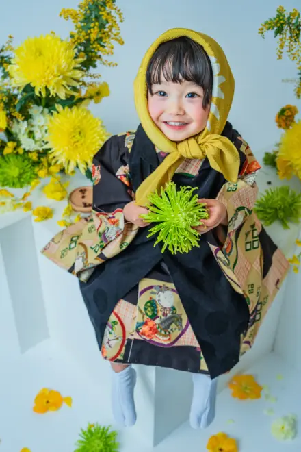 からし色のずきんと黒い絵柄着物を着て緑の菊を両手で抱え、黄色い花々に囲まれて笑顔で立つ七五三3歳の子ども