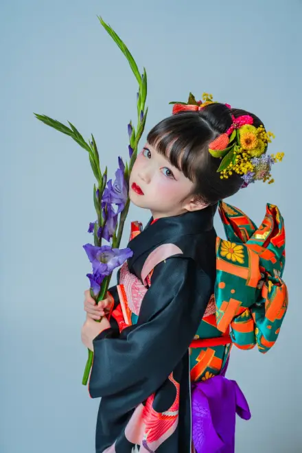 黒地の着物に色とりどりの花の髪飾りをつけた子どもが、紫のアイリスを抱えて横顔を見せる