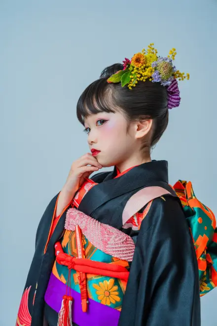 黒地の着物に黄色いミモザと生花の髪飾りをつけた子どもが、横顔を見せながら顎に手を添える