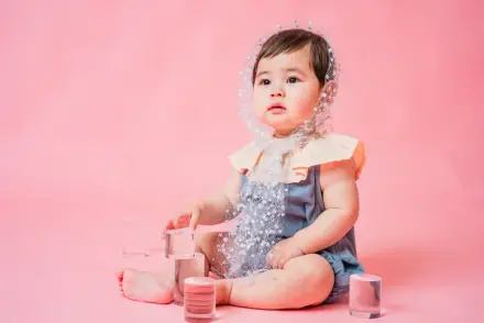 青いレースロンパースとビーズのヘッドドレスを着た赤ちゃんがピンク背景でキューブブロックを触りながら座る