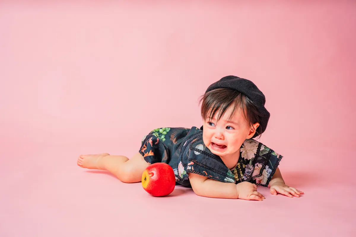 ピンク背景でベレー帽をかぶり林檎とうつ伏せになる赤ちゃん(Classic Portrait シーン)
