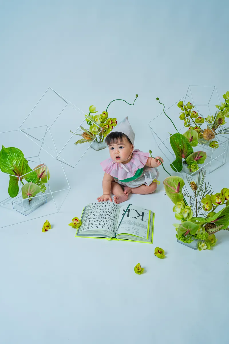 開いた本と緑の花に囲まれて座る赤ちゃん(Garden シーン)